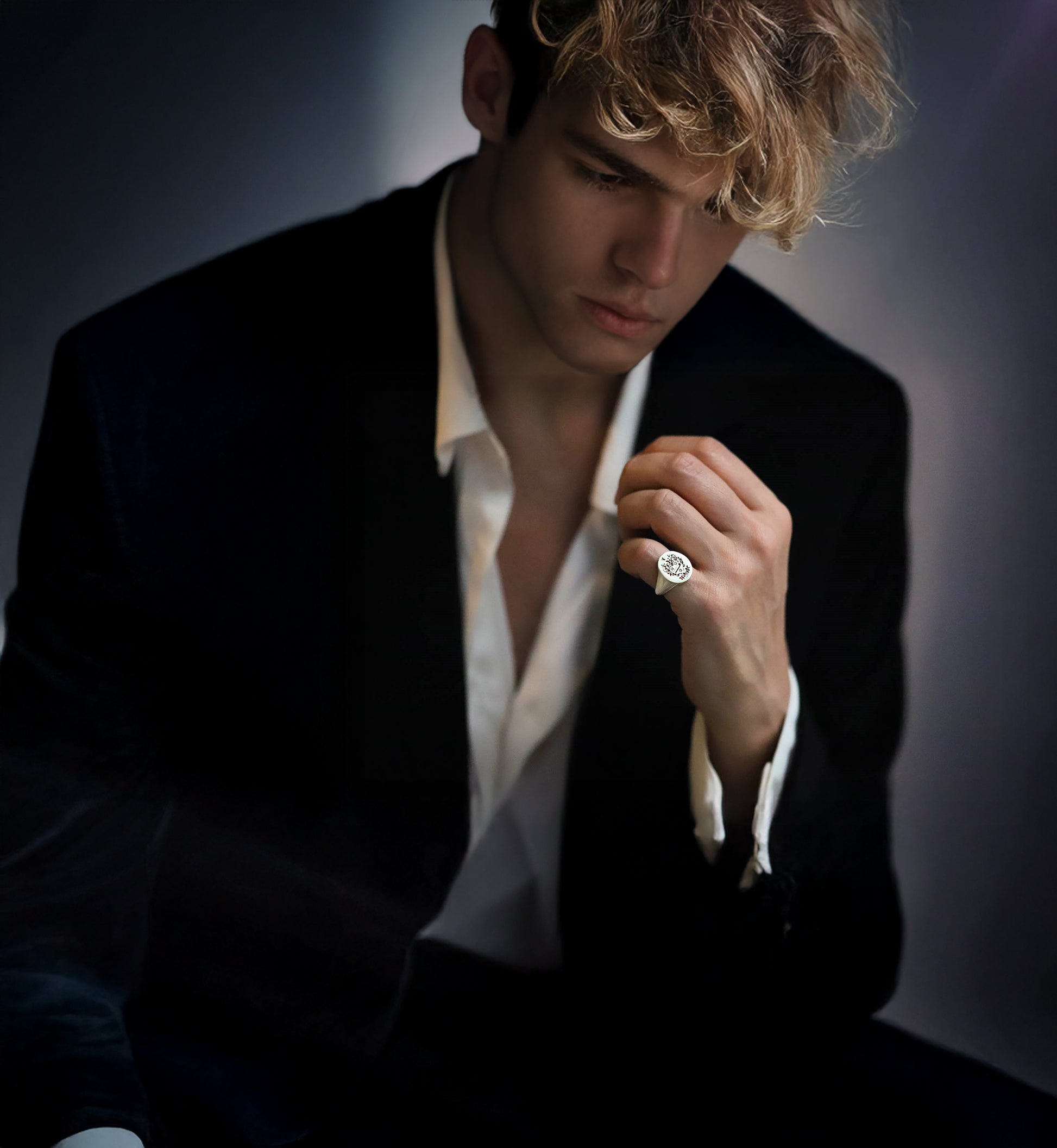 Man wearing a gold pinky signet ring, black blazer and white against a dark background