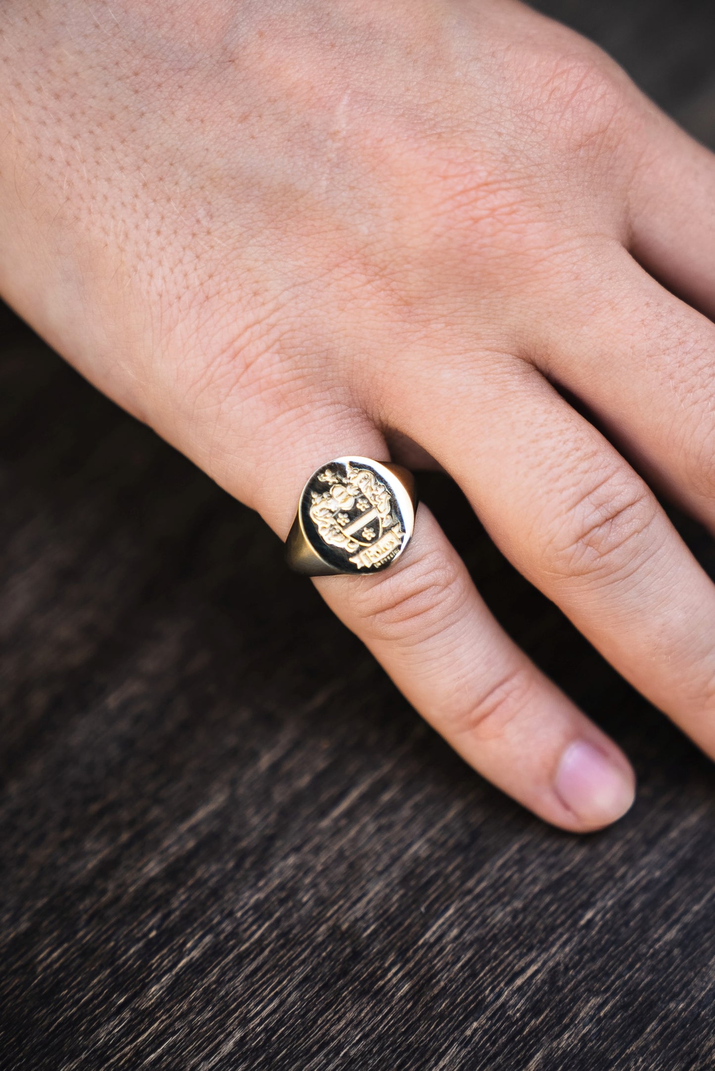 solid gold signet ring with a family crest design on a men's pinky finger against a dark wooden background