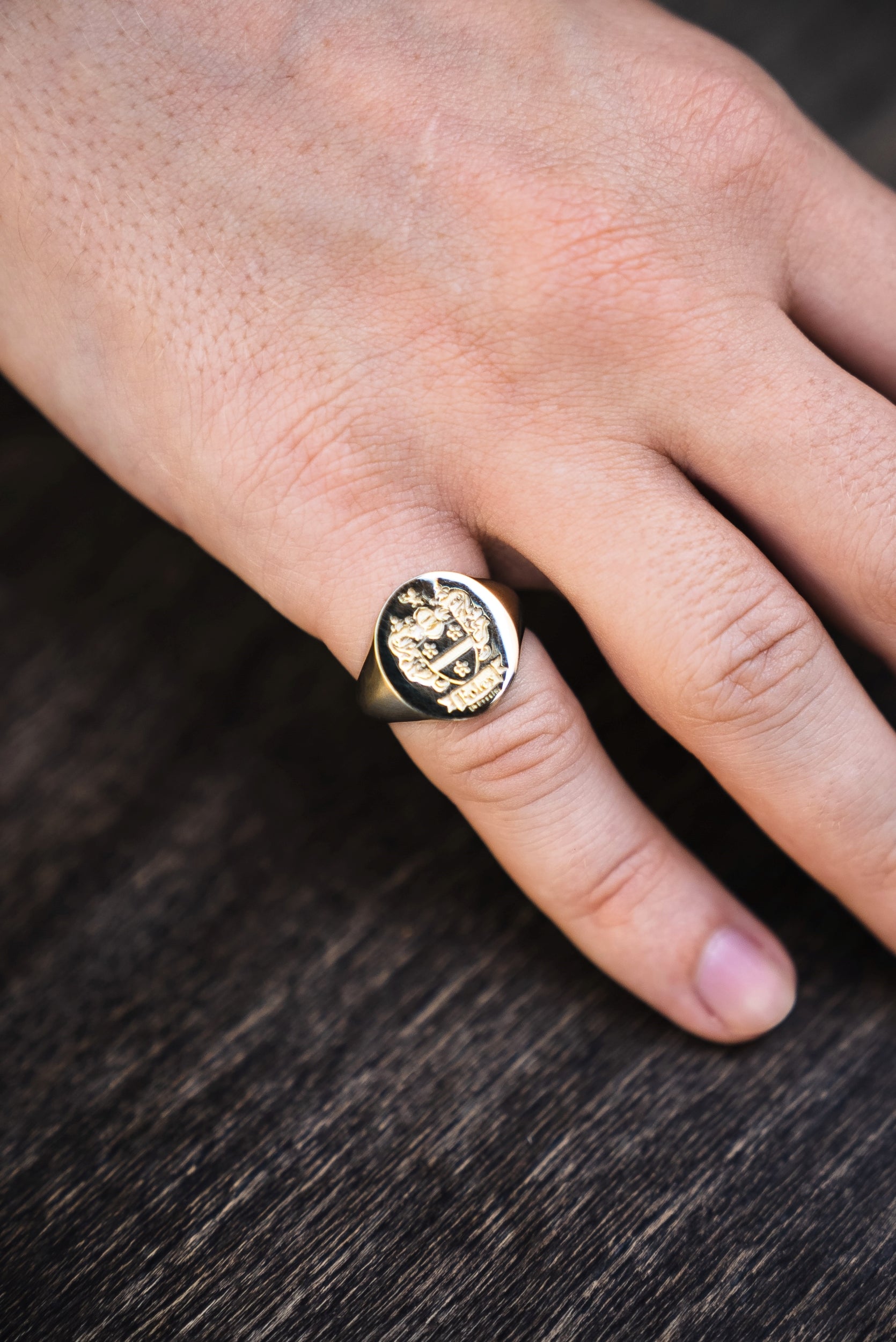 solid gold signet ring with a family crest design on a men's pinky finger against a dark wooden background