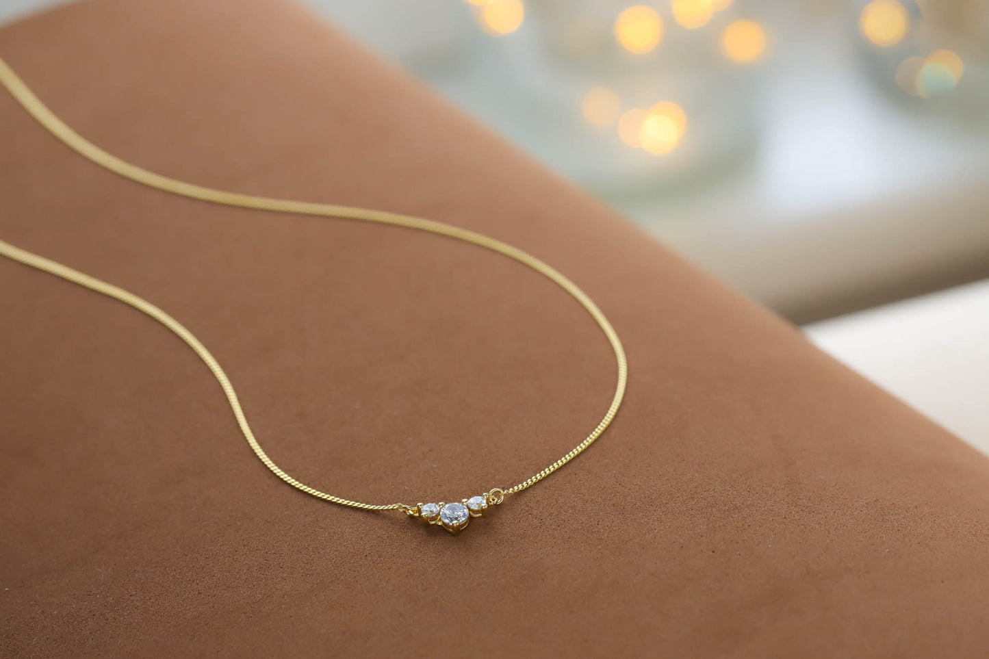 Close up of the delicate gold chain and 3 stone cz pendant resting on a textured brown surface.