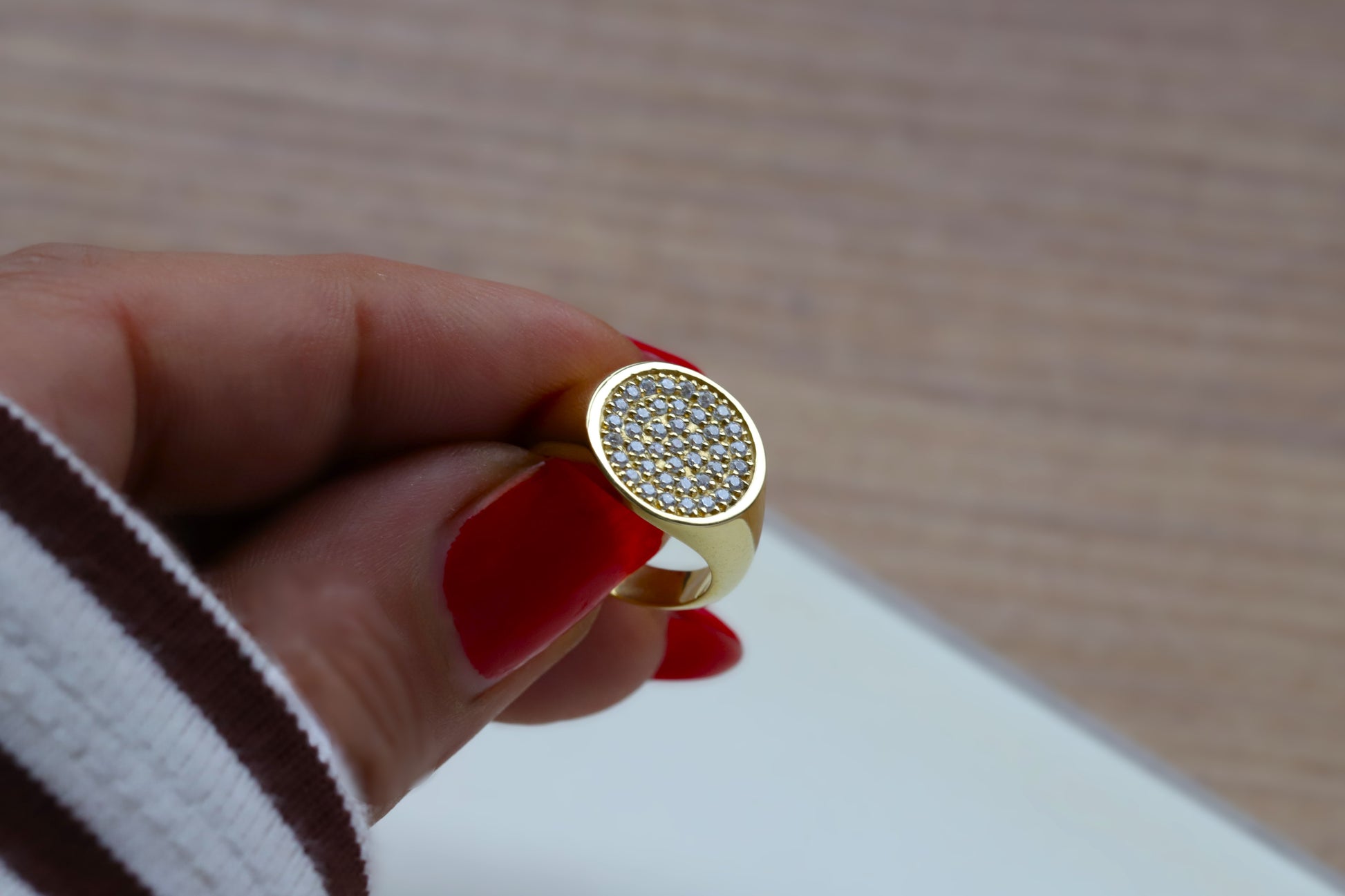 Gold signet ring with clear cz gemstones held by a hand with red nail polish on a wooden surface. 