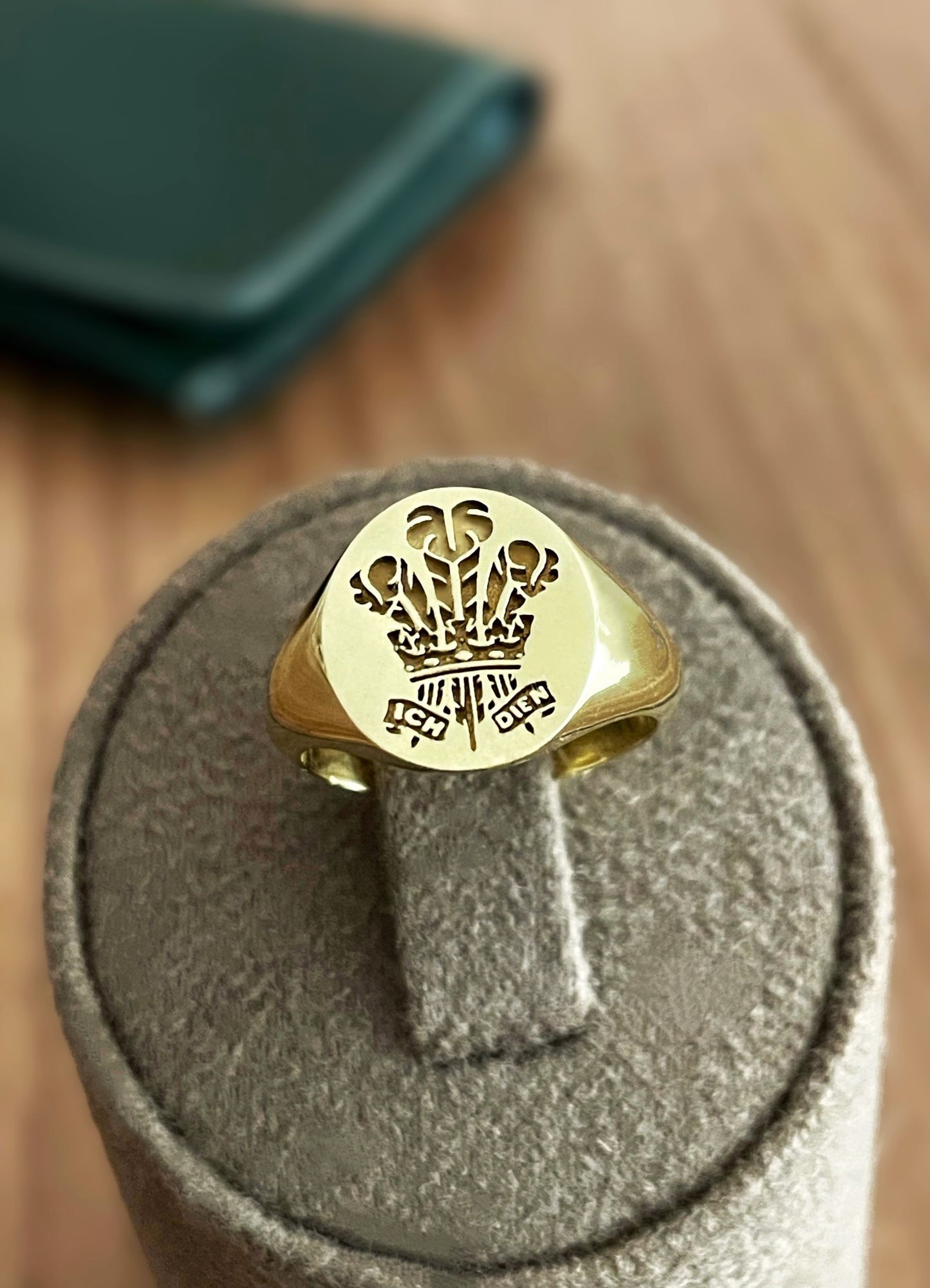 Gold signet ring engraved with a custom crown design with a blurred green object in the background.