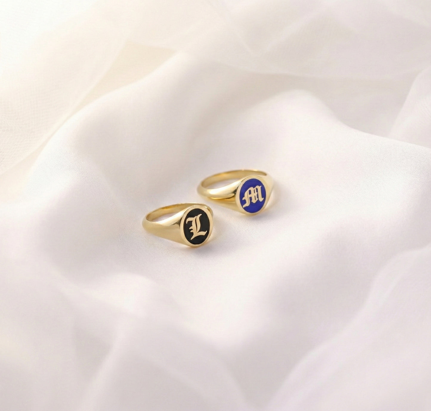 gold initial signet rings with blue and black enamel on a white fabric background