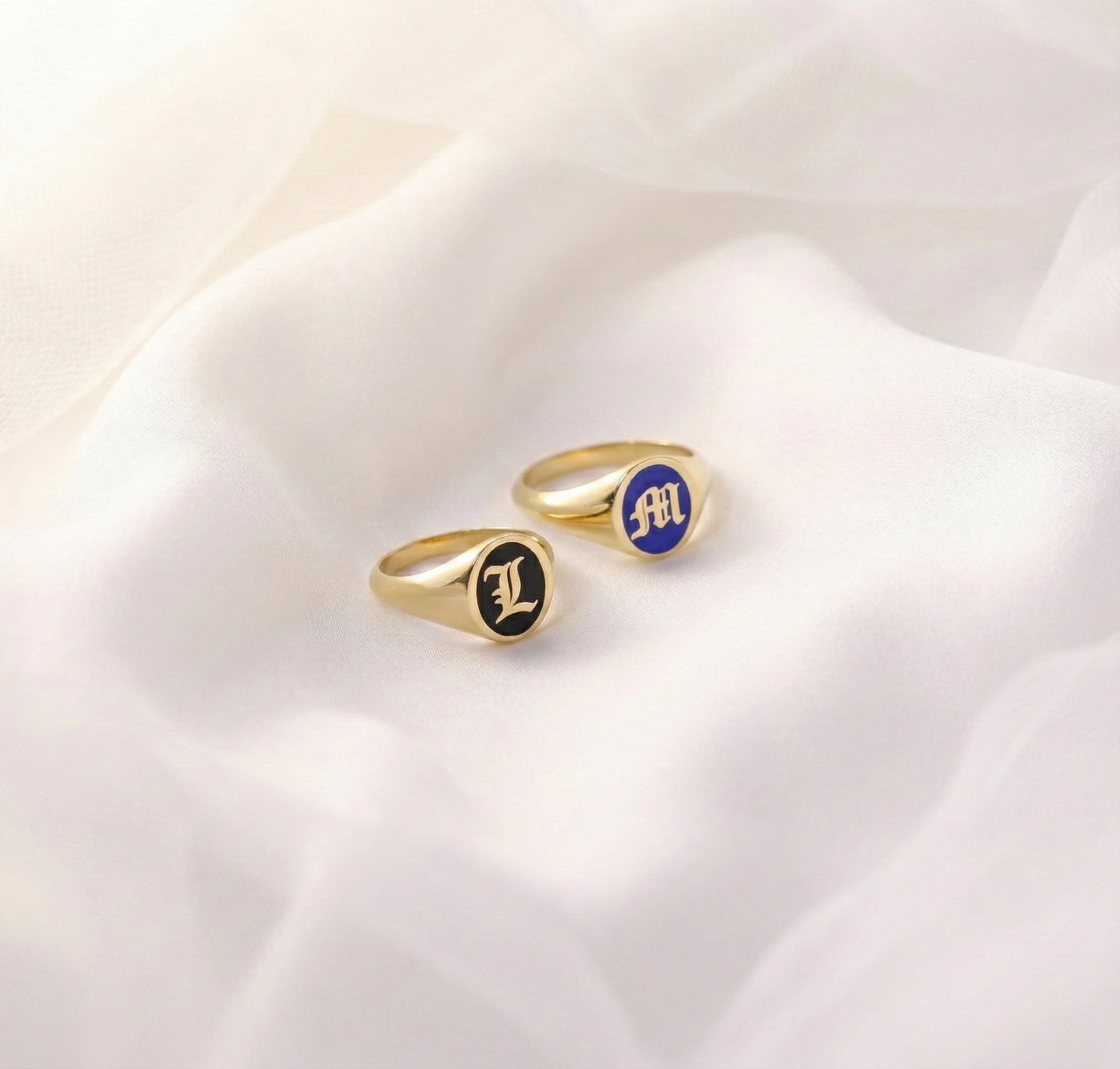 gold initial signet rings with blue and black enamel on a white fabric background