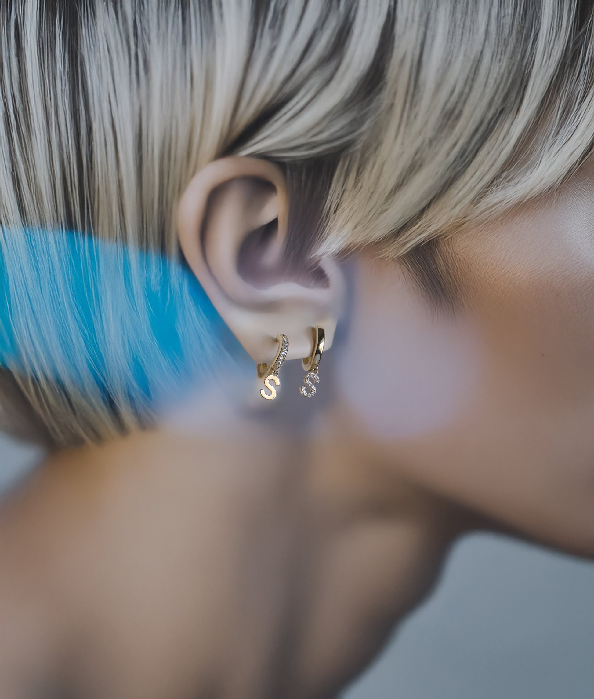 Close-up of a model wearing gold letter huggie hoop earrings set with a blurred background