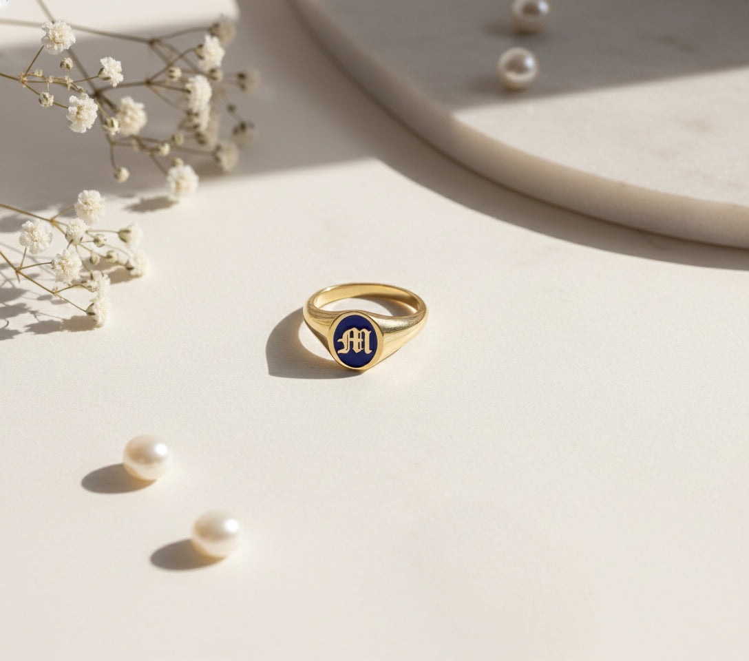 Gold initial signet ring with blue enamel on a light surface with pearls and flowers